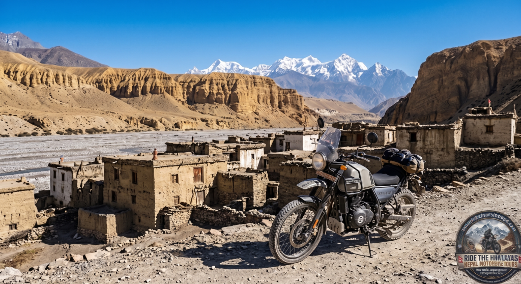 A single classic-styled adventure motorcycle with luggage is parked on a dusty dirt trail. In the background are ancient mud-brick and stone buildings of Kagbeni village, immense sculpted canyon cliffs, and distant snow-capped Himalayan peaks under a clear blue sky. A detailed tour logo is in the bottom right corner.
